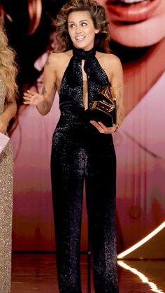 a woman standing on top of a stage wearing a black jumpsuit and holding an award