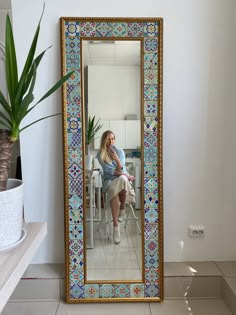 a woman sitting on a chair in front of a mirror that is next to a potted plant