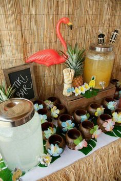 a table topped with pineapples and cups filled with liquid next to a flamingo