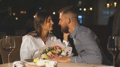 a man and woman sitting at a table with flowers in front of each other, looking into each others eyes