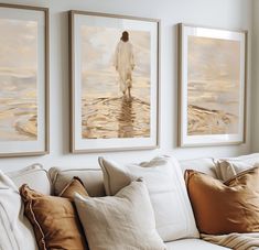 three framed paintings hang on the wall above a white couch with pillows and throw pillows