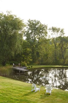 two lawn chairs sitting on top of a lush green field next to a lake and dock