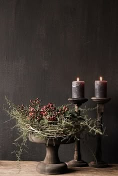 two candles sitting on top of a table next to a bowl filled with flowers and greenery