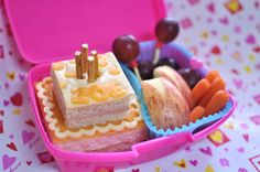 a pink container filled with cake and fruit on top of a table next to other food items