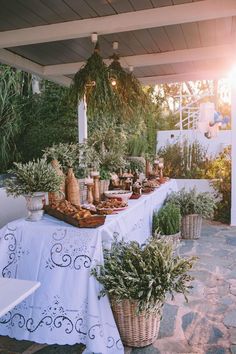 an outdoor buffet with bread and vegetables on the table, surrounded by greenery as well as potted plants