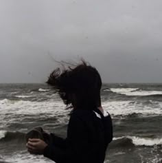 a person standing in front of the ocean on a cloudy day with their hair blowing in the wind