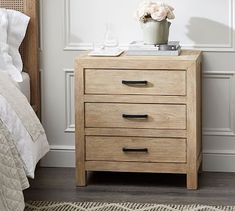 a nightstand with two drawers and a flower pot on it next to a white bed