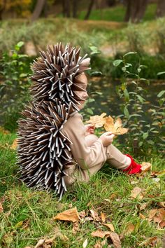 a small child sitting on the ground next to a hedgehog with leaves in it's mouth