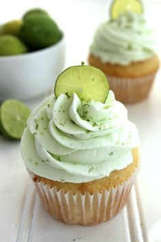 two cupcakes with white frosting and lime slices on the side next to a bowl of limes