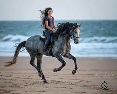 a woman riding on the back of a gray horse down a beach next to the ocean