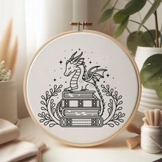 a cross stitch pattern with a dragon sitting on top of a stack of books in front of a potted plant