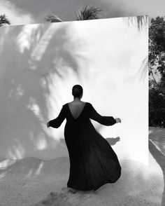 a woman in a long black dress standing next to a white wall and palm trees