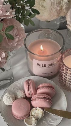 pink macaroons are on a plate next to a candle and vase with flowers