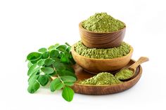 three wooden bowls filled with green powder next to a leafy plant on a white background