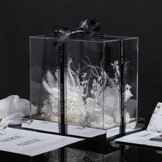 two clear boxes with white flowers in them on a black table next to some papers