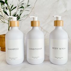 three body wash bottles sitting on top of a counter next to a potted plant