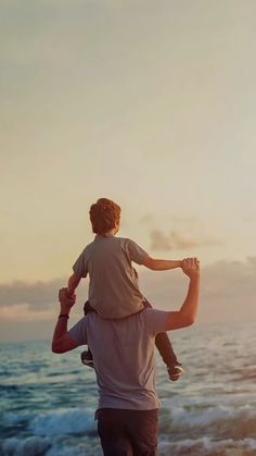 a man carrying a young boy on his shoulders while standing on the beach at sunset