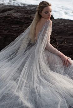 a woman sitting on top of a rock near the ocean wearing a long white dress