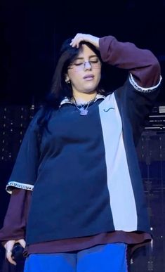 a woman standing on stage with her hands behind her head, wearing glasses and blue pants