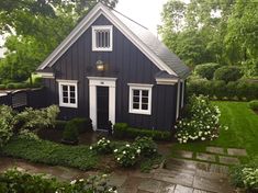 a small black house surrounded by lush green trees and bushes with white trim on the windows