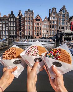 three people are holding up their food in front of some buildings on the water with boats in the background