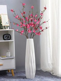 a white vase filled with pink flowers on top of a blue rug next to a book shelf