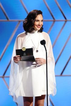 a woman standing at a podium with a microphone in her hand and wearing a white dress