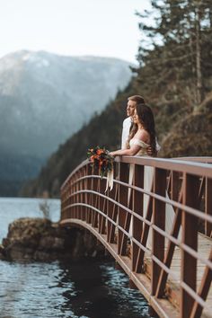 two people standing on a bridge over water with mountains in the background