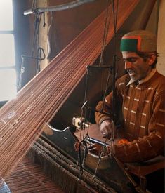 a man is weaving fabric on an old loom while wearing a green and red hat