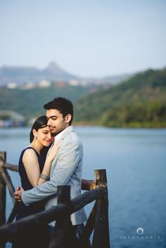 a man and woman standing next to each other on a bridge near the water with mountains in the background