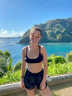 a woman standing on the side of a road next to water and mountains with trees in the background