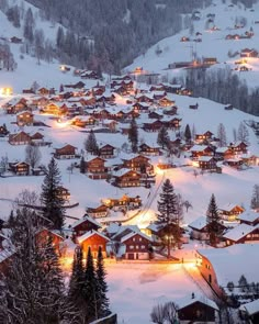 a village is lit up at night in the snow with trees and mountains behind it