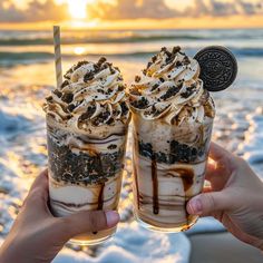 two people holding up glasses with ice cream and oreo cookies on them at the beach