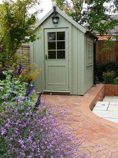 a garden shed with purple flowers in the foreground and a brick path leading to it