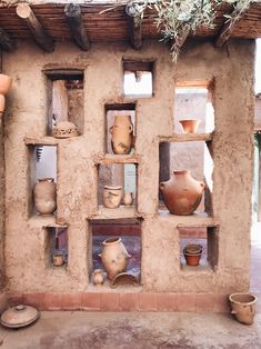 several clay pots are displayed on shelves in an adobe - style building, with plants hanging from the roof