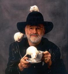 an old man wearing a black hat and holding two white kittens on his shoulders