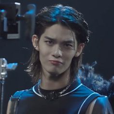 a young man with long hair wearing a black shirt and silver bracelets, standing in front of a dark background