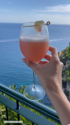 a person holding up a wine glass with a drink in it on a balcony overlooking the ocean