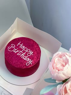 a pink cake with happy birthday written on it in a white box next to flowers