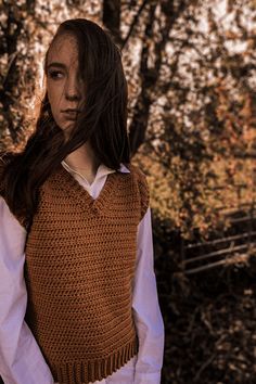 a woman standing in front of trees wearing a brown sweater and white shirt with long sleeves