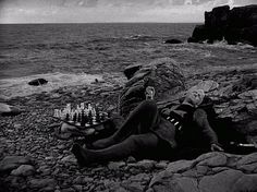 a woman is laying on the rocks with chess pieces in her lap and looking out at the ocean