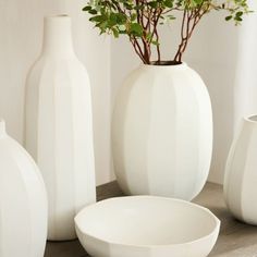 three white vases and one bowl on a table with flowers in them, all sitting next to each other