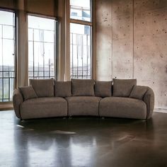 a large sectional couch in an empty room with two windows and concrete walls behind it