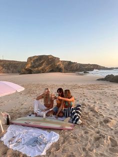 four people are sitting on the beach with their surfboards in front of an umbrella