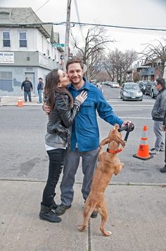 a man and woman standing next to a brown dog on a sidewalk with an orange cone in front of them