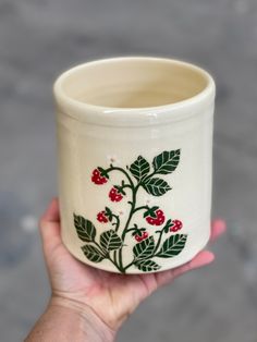 a hand holding a white cup with red berries and leaves painted on the outside of it