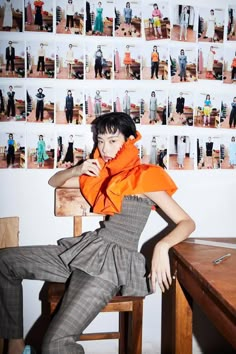 a woman in an orange top and plaid pants sitting at a desk with pictures on the wall behind her