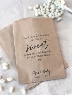 three bags with wedding favors on them sitting next to some white flowers and beans in the background