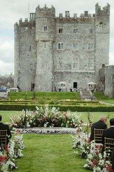 two men sitting on chairs in front of an old castle with flowers and greenery