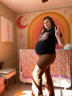 a pregnant woman standing in front of a bed giving the peace sign